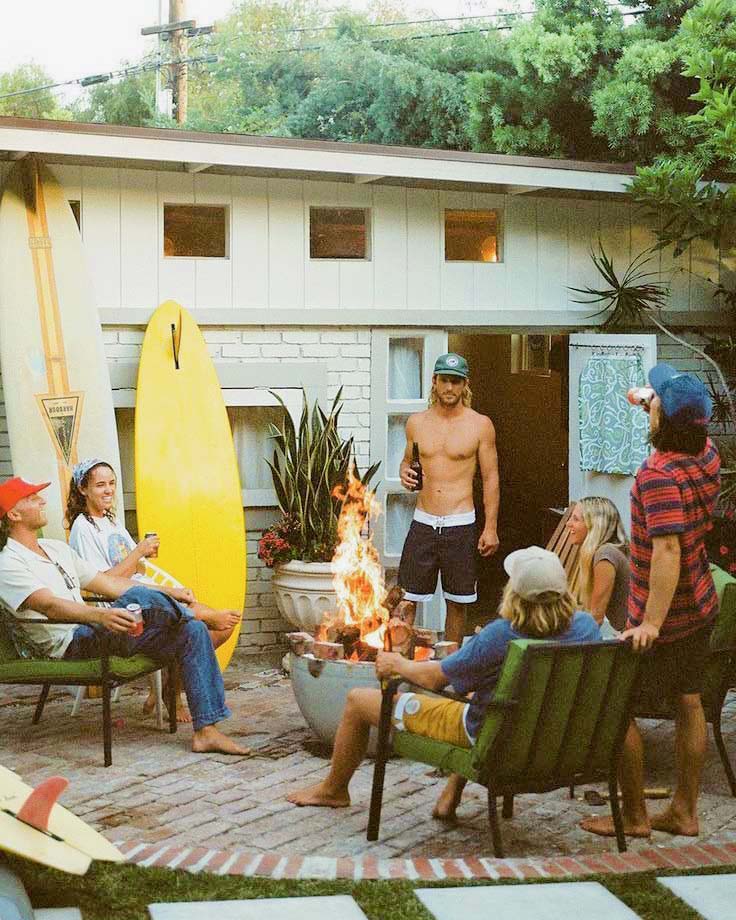 Group of people gathered around a fire pit in a backyard wearing Vintage Surfewar Clothing with a surfboard and house in the background.