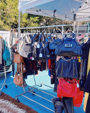 Display of handbags at an outdoor market with a blue tarp and trees in the background.