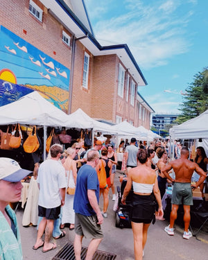 Outdoor market scene with people shopping under white tents against a building with a mural.