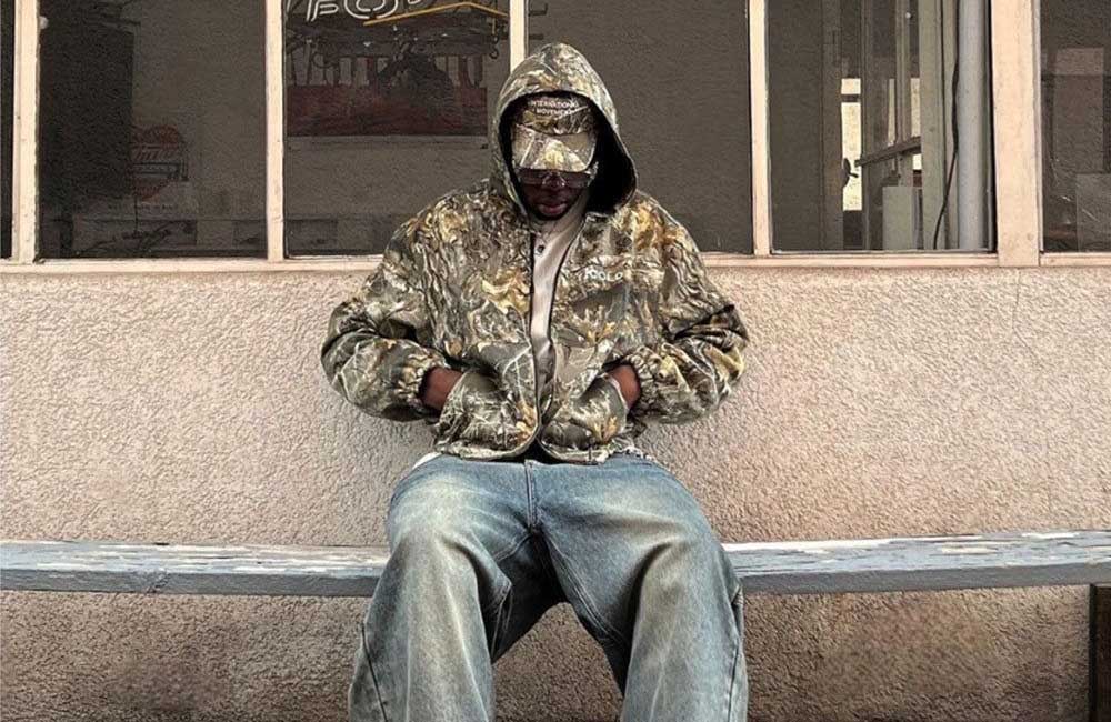Person wearing a vintage Realtree camouflage jacket sitting on a bench in front of a building.