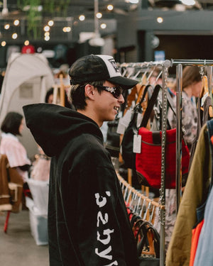 Person wearing a black hoodie with white text, standing in an indoor vintage market with bags on display.