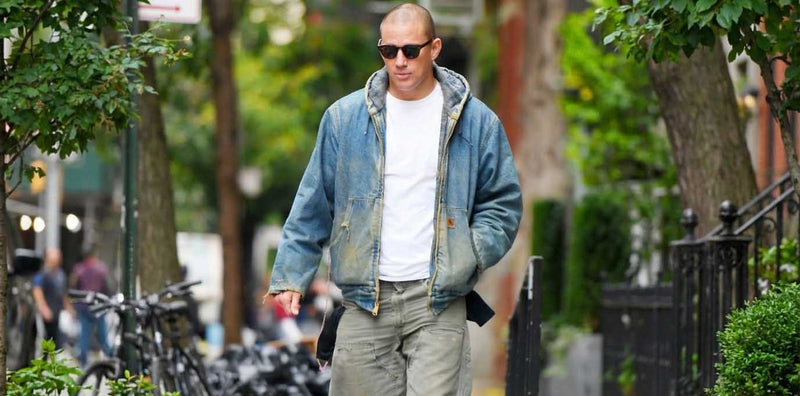 Man walking outdoors on a city street with trees and bicycles in the background wearing a distressed vintage Carhartt jacket