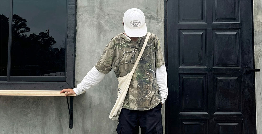 Person wearing a vintage Realtree camouflage jacket and white cap leaning against a building with a black door.