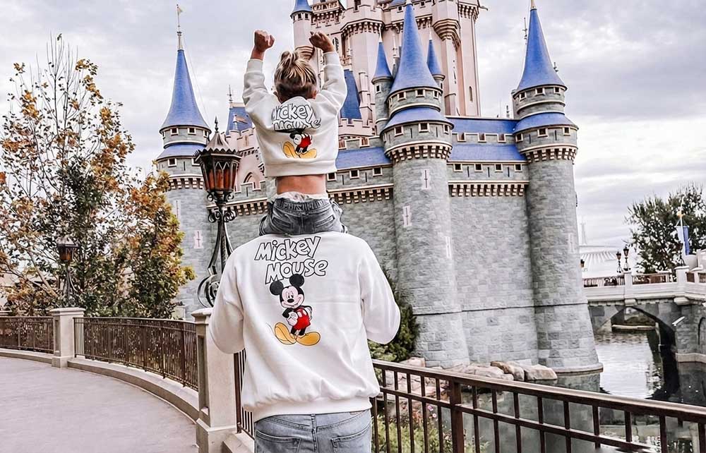 Person wearing a vintage Mickey Mouse sweatshirt in front of a castle, likely at Disneyland.