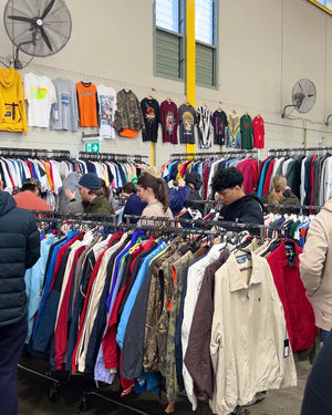 People shopping in a vintage clothing market store with racks of clothes and fans on the wall.