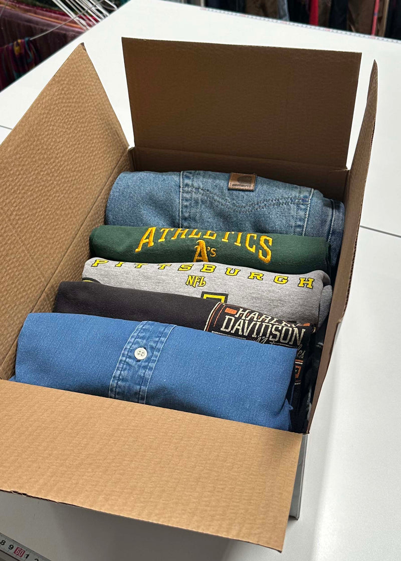 Vintage Mystery Box containing folded Carhartt  jeans, Oakland Athletics T-Shirt, Bittsburgh Steelers Sweatshirt, Harley Davidson T-Shirt, and Polo Button Up Shirt on a white bench in a warehouse