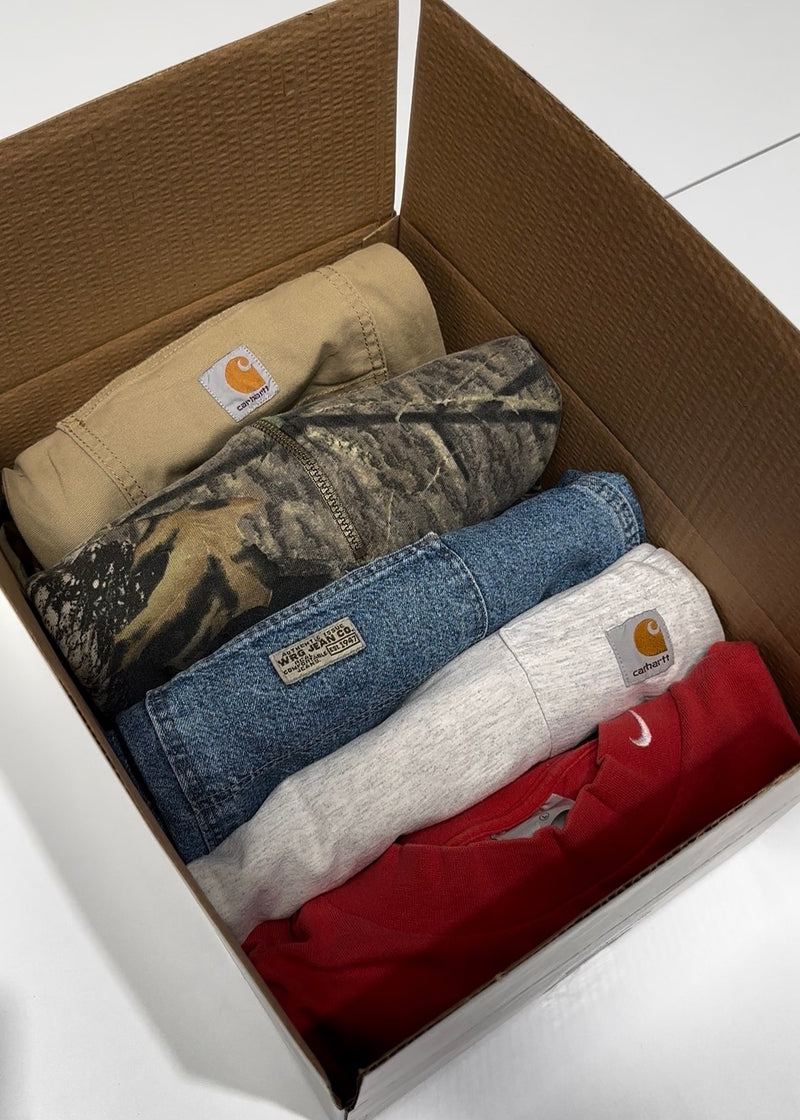 Vintage Mystery Box containing folded Carhartt T-Shirt, Carhartt Pants, Wrangler Denim Carpenter Shorts, Nike Swoosh T-Shirt, and Realtree Camo Quarter Zip Sweatshirt on a white bench in a warehouse