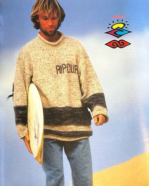 Man wearing a sweater with 'Rip Curl' branding, holding a surfboard against a blue sky and sand background.