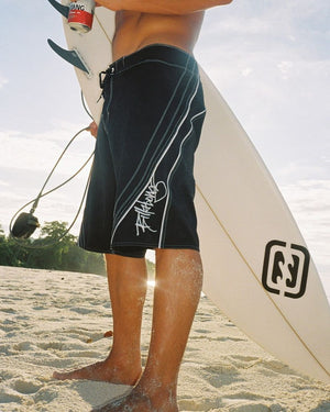 Person holding a surfboard on a beach wearing black Billabong board shorts with white designs.