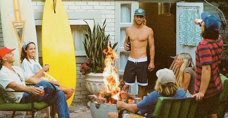 Group of people gathered around a fire pit in a backyard wearing Vintage Surfewar Clothing with a surfboard and house in the background.