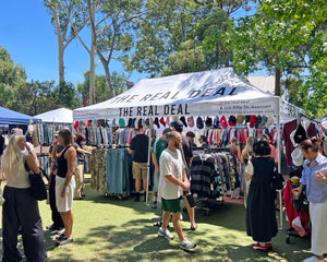 Outdoor vintage market scene with 'The Real Deal' clothing stall under a white tent.