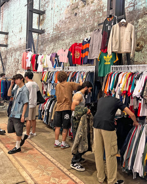 People browsing through a vintage clothing stall with racks of clothes against a brick wall.
